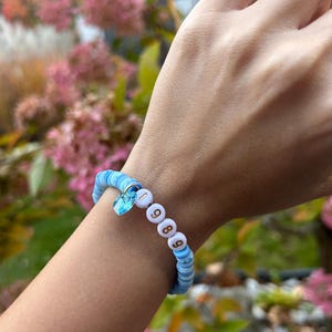 May include: A blue and white beaded bracelet with a small blue crystal charm and gold beads spelling out "1989".