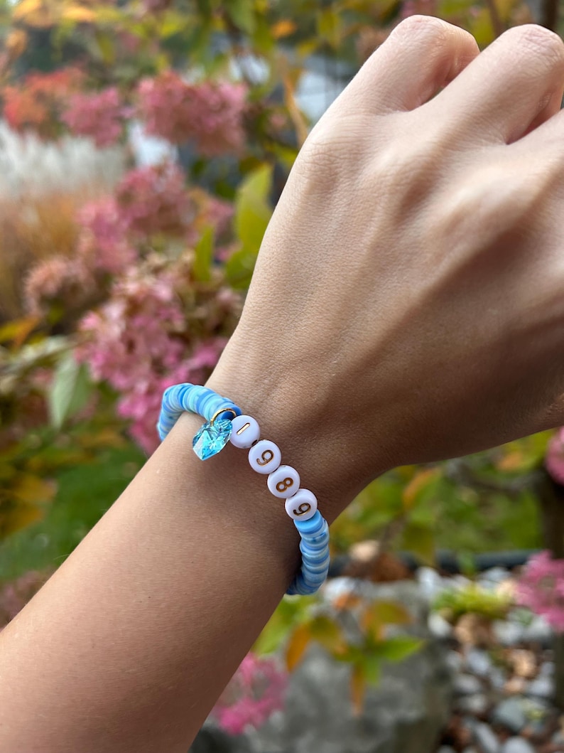 May include: A blue and white beaded bracelet with a small blue crystal charm and gold beads spelling out "1989".