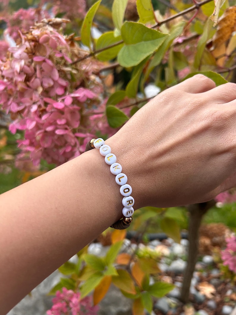 May include: A white beaded bracelet with gold lettering spelling out the word "FOLKLORE".