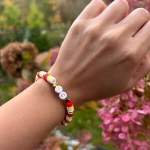 May include: A white, red, and yellow beaded bracelet with gold star and letter charms. The bracelet is on a person's wrist.