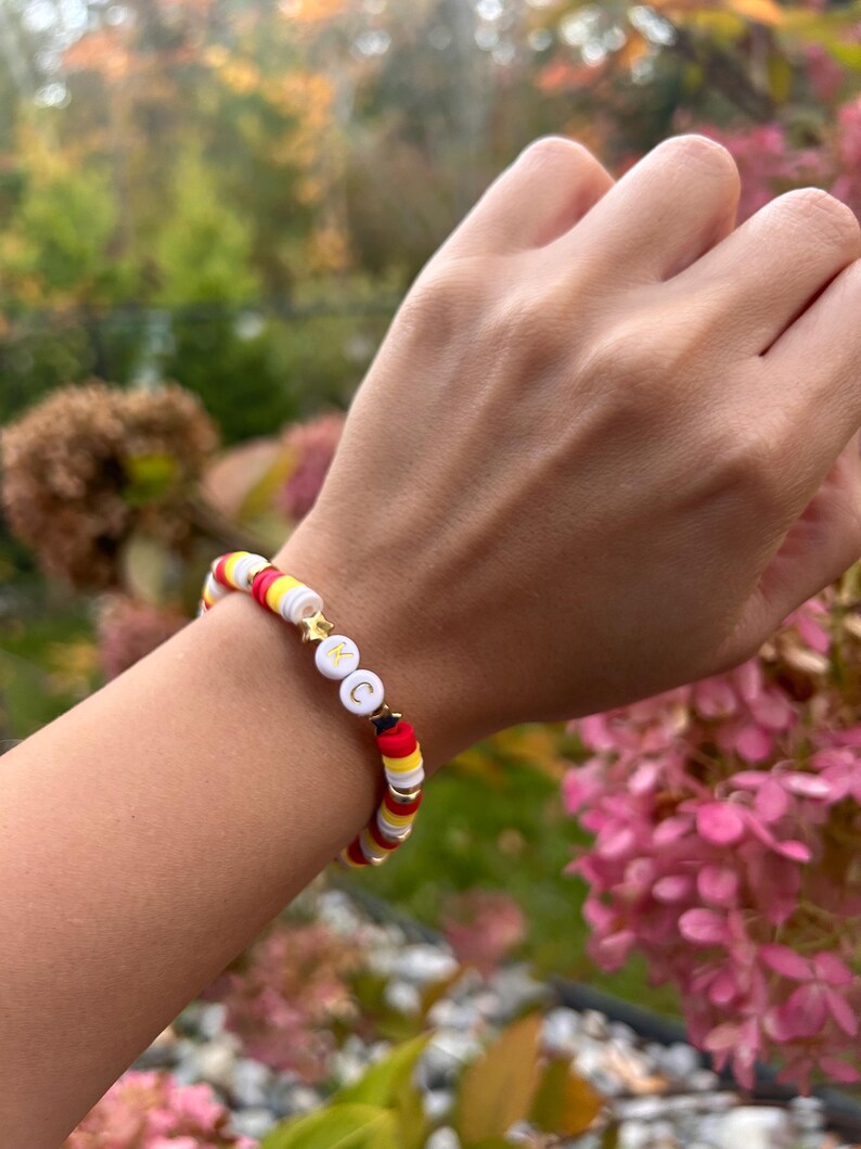 May include: A white, red, and yellow beaded bracelet with gold star and letter charms. The bracelet is on a person's wrist.