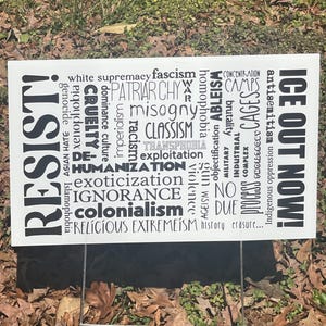 May include: A white sign with black text, including the words "RESIST!" and "ICE OUT NOW!" Various terms like "racism," "misogyny," and "colonialism" are also displayed. The sign is supported by a metal frame.