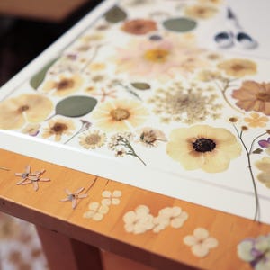 May include: A white rectangular board covered in an arrangement of pressed dried flowers in shades of cream, yellow, and muted orange. The board rests on a light wood table surface that is also decorated with several small individual pressed flower petals.