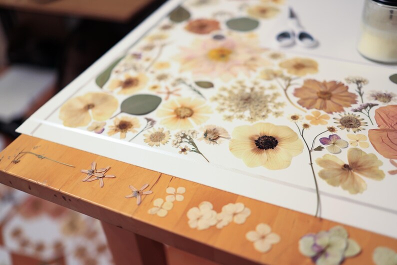 May include: A white rectangular board covered in an arrangement of pressed dried flowers in shades of cream, yellow, and muted orange. The board rests on a light wood table surface that is also decorated with several small individual pressed flower petals.