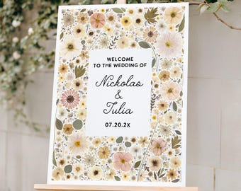 May include: A rectangular wedding welcome sign on a wooden easel featuring a border of pressed flowers in shades of yellow, cream, and muted pink. The center text reads: WELCOME TO THE WEDDING OF Nickolas & Tulia 07.20.2X in black lettering.