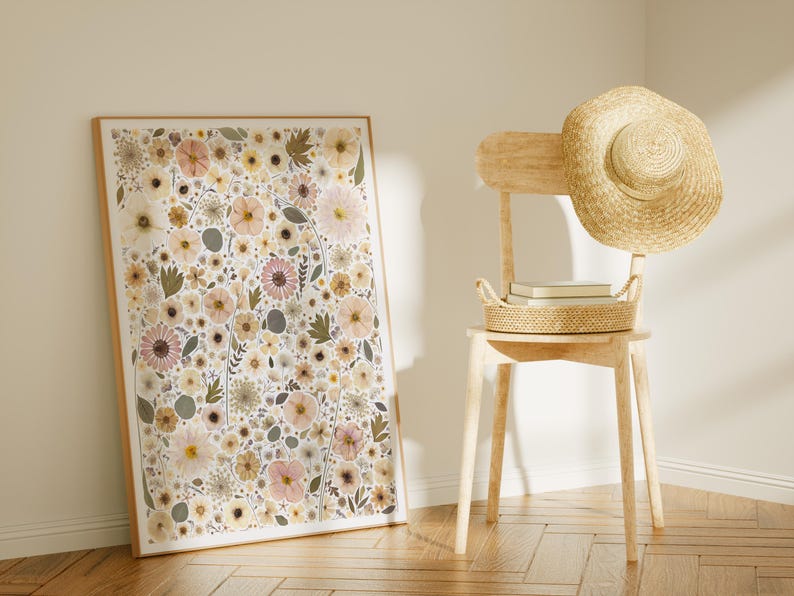 May include: A rectangular framed wall art print featuring a dense botanical pattern of pressed-style flowers in shades of pale pink, cream, muted yellow, and sage green, displayed next to a light-colored wooden chair with a straw hat and basket.