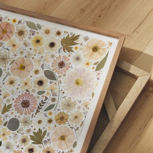 May include: A framed wall art piece featuring a collection of pressed flowers and leaves in shades of cream, pale yellow, soft pink, and muted green. The botanical arrangement is displayed on a white background within a light wood frame.