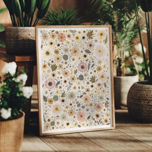 May include: A rectangular framed print featuring a dense collage of pressed flowers and foliage in shades of cream, pale yellow, soft pink, and muted green. The artwork is encased in a light-toned wooden frame with a clean, simple border design.