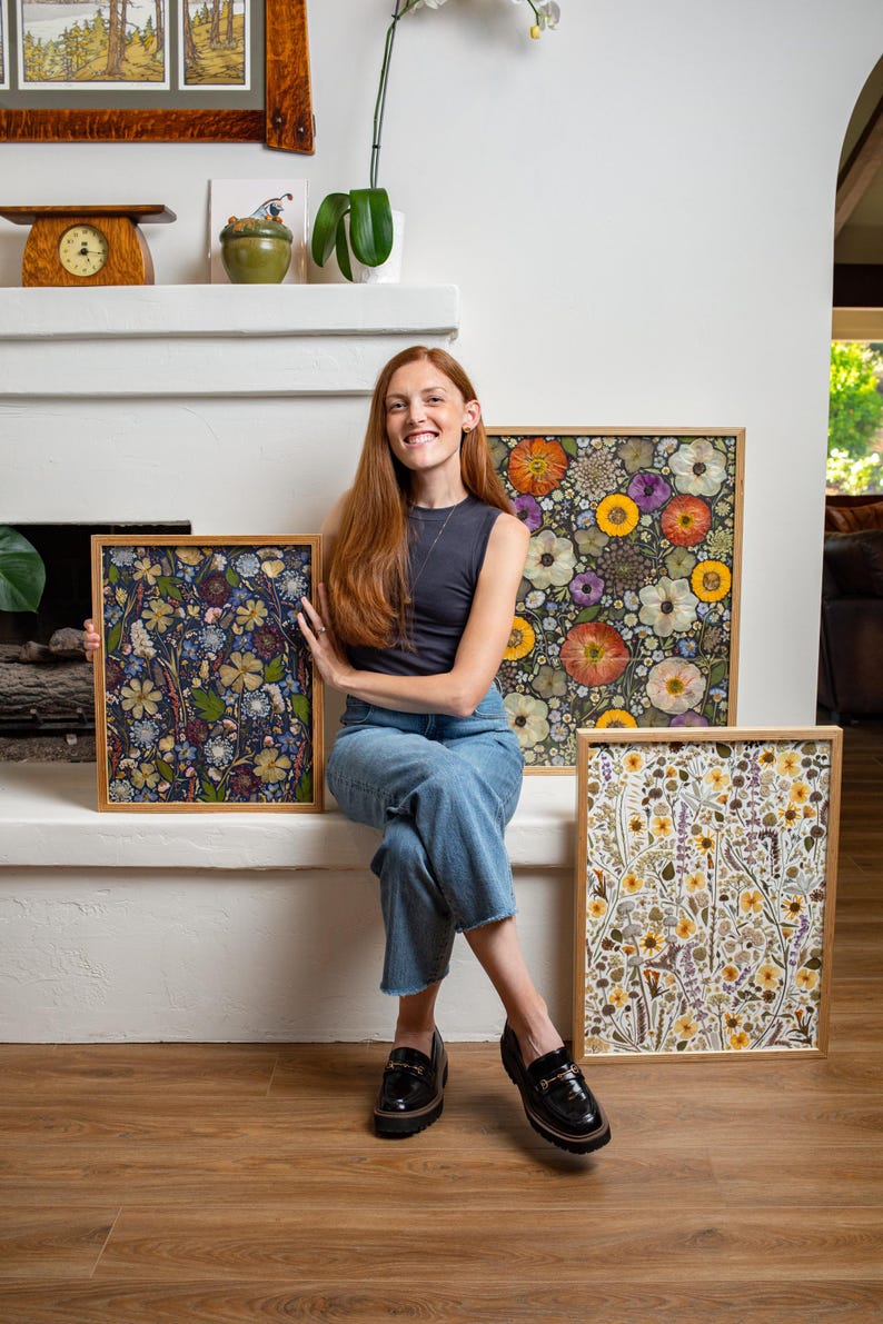 May include: Three rectangular framed art pieces featuring pressed flowers. One frame displays a dark blue background with yellow and white blossoms. Another shows vibrant orange, yellow, and purple flowers. The third features a light, botanical arrangement.