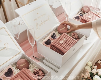 May include: White gift boxes with gold lettering that reads "Will You Be My Bridesmaid?" are open to reveal pink accessories. Items include heart-shaped sunglasses, cosmetic bags, and scrunchies. The boxes are arranged on a white table with champagne flutes.