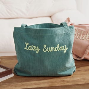 May include: A teal corduroy tote bag with the embroidered text "Lazy Sunday" in a light green script. The bag has two handles and is sitting on a wooden surface. A pink tote bag with the word "Bride" is partially visible.