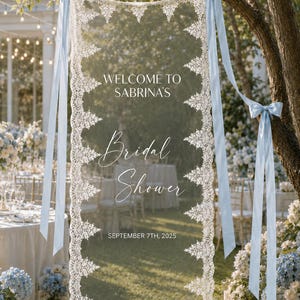 Op de afbeelding: Een rechthoekig transparant bord met witte kanten randen langs de verticale zijden en lichtblauwe satijnen linten aan de bovenhoeken. De tekst luidt: WELCOME TO SABRINA'S Bridal Shower SEPTEMBER 7TH, 2025 in witte letters.