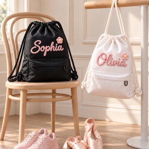 Puede incluir: Una mochila de cordón negra con el nombre Sophia y una pequeña flor rosa, y una mochila de cordón blanca con el nombre Olivia y una pequeña flor rosa. Ambas bolsas tienen un bolsillo frontal con cremallera y correas de cuerda gruesa.