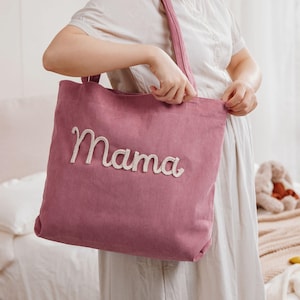 May include: A dusty rose corduroy tote bag with the word "Mama" embroidered in a cream-colored script. The bag has a matching corduroy strap and is held by a person wearing a white dress.