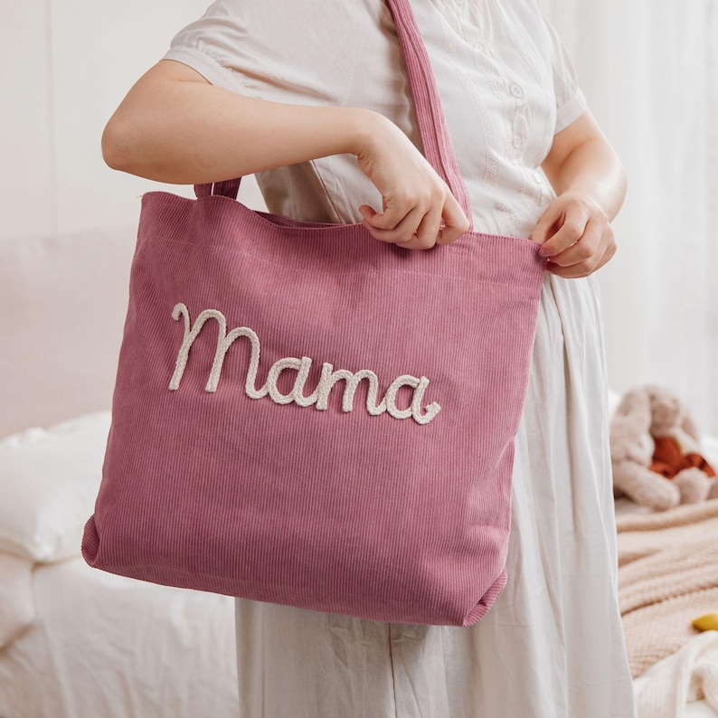 May include: A dusty rose corduroy tote bag with the word "Mama" embroidered in a cream-colored script. The bag has a matching corduroy strap and is held by a person wearing a white dress.