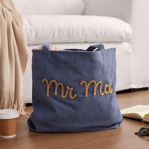 May include: A blue corduroy tote bag with the embroidered words "Mr Ma" in gold thread. The bag has two handles and is sitting on a wooden floor. A white couch is in the background.