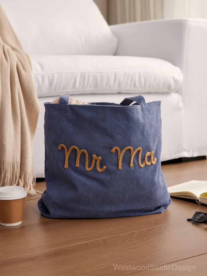 May include: A blue corduroy tote bag with the embroidered words "Mr Ma" in gold thread. The bag has two handles and is sitting on a wooden floor. A white couch is in the background.