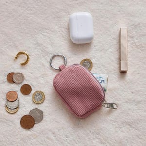 May include: A flat lay featuring a pink corduroy coin purse with a silver zipper, a gold hoop earring, a white wireless earbud case, a tube of lipstick, and various coins. The items are arranged on a white, textured surface.