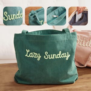 May include: A green corduroy tote bag with the words "Lazy Sunday" embroidered in a light green script. The bag has two handles and a zippered pocket. A close-up shows the word "Sunday" on the bag.