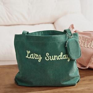 May include: A dark green corduroy tote bag featuring the text Lazy Sunday embroidered in light green thread. The bag includes two shoulder straps and a small matching corduroy zippered pouch attached to a silver metal ring on the side.