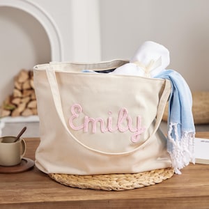 May include: A beige corduroy tote bag with the name "Emily" embroidered in pink. The bag contains a rolled white towel tied with a ribbon and a blue and white fringed towel. The bag is on a woven mat.