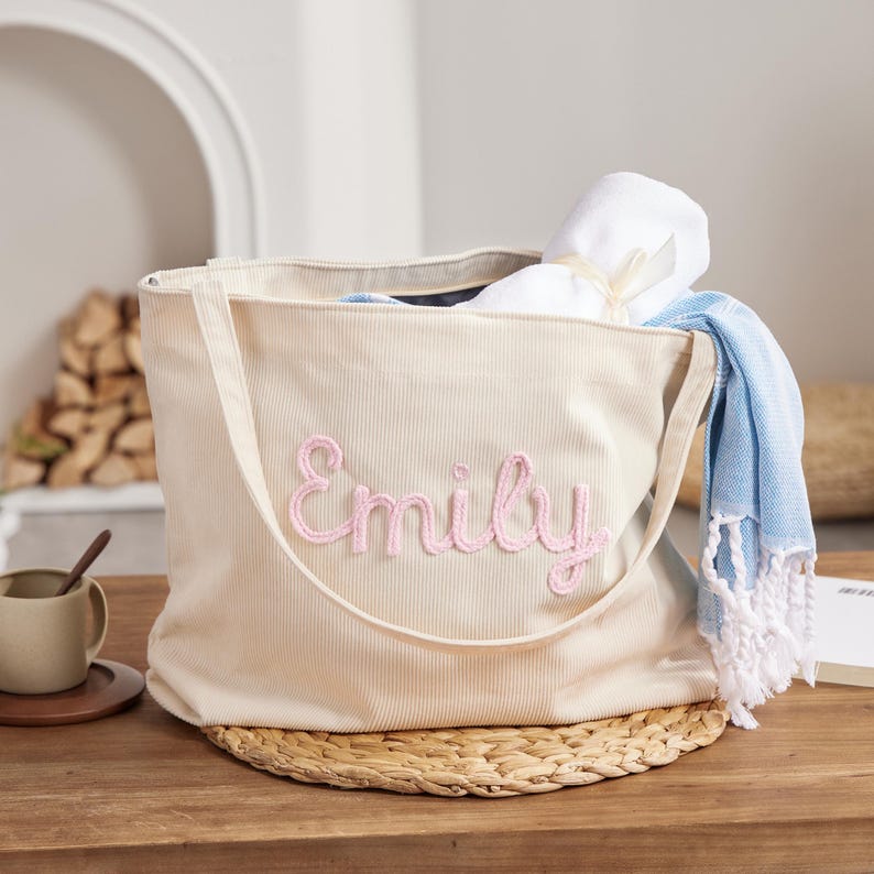 May include: A beige corduroy tote bag with the name "Emily" embroidered in pink. The bag contains a rolled white towel tied with a ribbon and a blue and white fringed towel. The bag is on a woven mat.