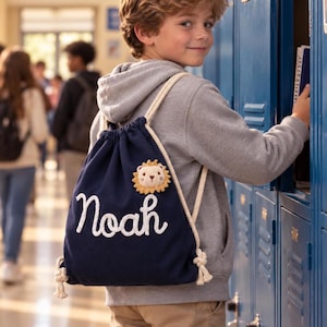 May include: A navy blue drawstring backpack featuring the name Noah in white cursive lettering and a small, plush, circular lion face patch with a tan mane. The bag is constructed from fabric and includes thick, cream-colored rope shoulder straps.
