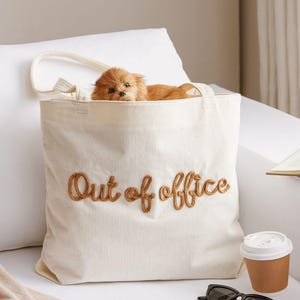 May include: Cream-colored corduroy tote bag with the embroidered phrase "Out of office" in brown thread. A small, fluffy, brown dog peeks out from the bag. Sunglasses and a coffee cup are next to the bag.