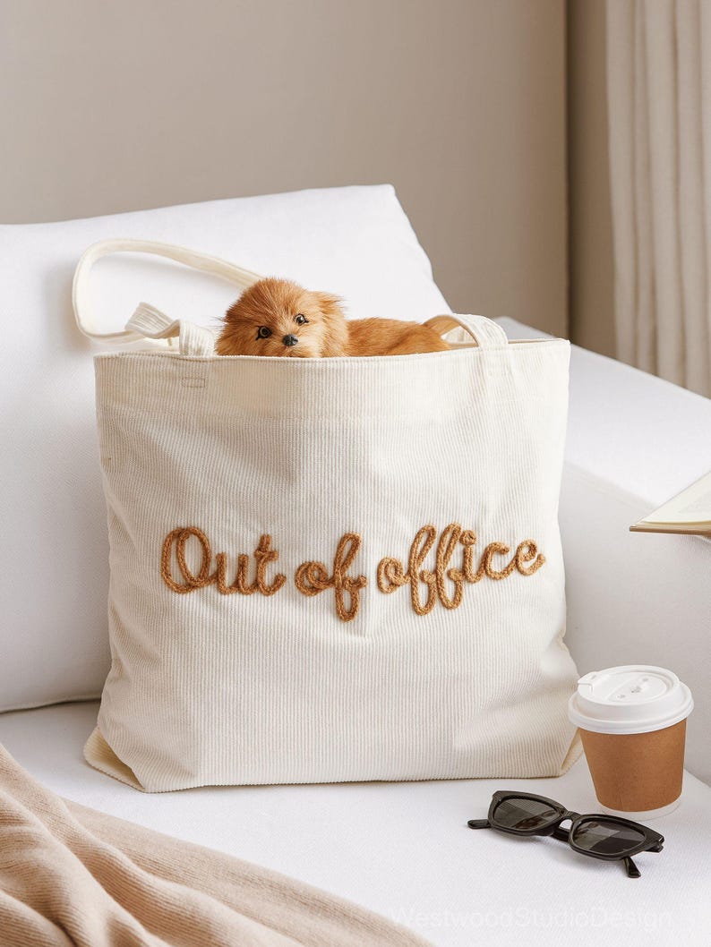 May include: Cream-colored corduroy tote bag with the embroidered phrase "Out of office" in brown thread. A small, fluffy, brown dog peeks out from the bag. Sunglasses and a coffee cup are next to the bag.