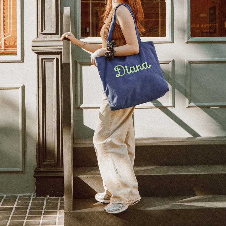 May include: A blue tote bag with the name "Diana" embroidered in green script. The bag has shoulder straps and is made of a soft fabric. The person is standing on steps in front of a door.