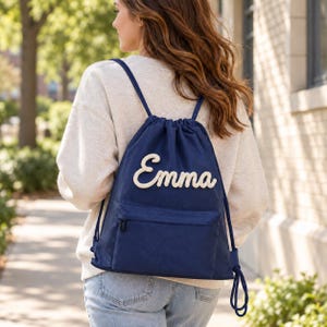 May include: A navy blue drawstring backpack featuring a front zippered pocket and the name Emma in white textured script lettering. The bag has matching navy blue cord straps and is worn over the shoulders with a light-colored sweatshirt.