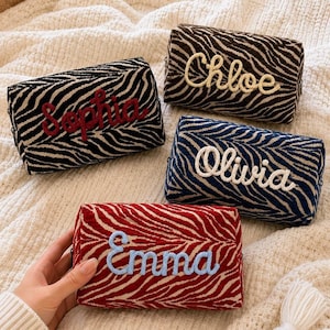 Può includere: Quattro astucci rettangolari con cerniera e un motivo a strisce zebrate testurizzato. Ogni astuccio presenta un nome ricamato in corsivo: Sophia in rosso, Chloe in crema, Olivia in bianco ed Emma in azzurro su sfondo rosso e crema.