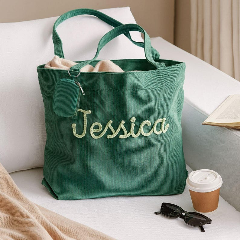 May include: A green corduroy tote bag with the name "Jessica" embroidered in a lighter green script. The bag has matching handles and a small attached coin purse. Sunglasses and a coffee cup sit nearby.