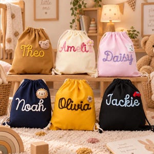 Puede incluir: Seis mochilas de cordón en color tostado, crema, rosa, azul marino, amarillo y negro. Cada bolsa presenta un nombre bordado en cursiva y un pequeño aplique de peluche, incluyendo un oso, una flor, un conejo, un león y un coche rojo.