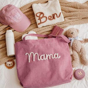 May include: A collection of baby and mom-themed items, including a pink corduroy tote bag with "Mama" embroidered on it, a pink baseball cap with "Mom Est. 2023", a white sweater with "Ben" on it, a teddy bear, and a white water bottle.