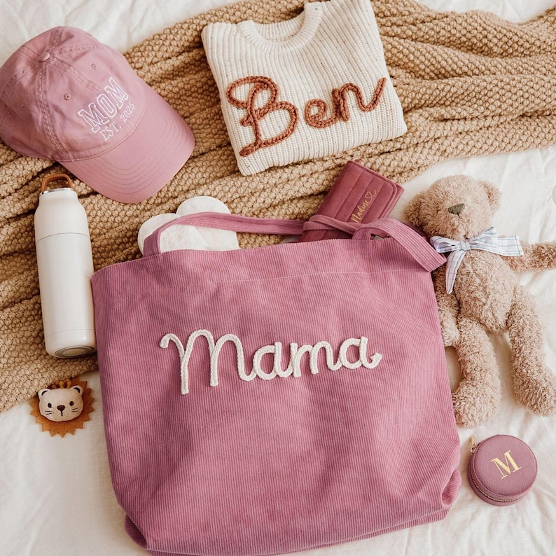 May include: A collection of baby and mom-themed items, including a pink corduroy tote bag with "Mama" embroidered on it, a pink baseball cap with "Mom Est. 2023", a white sweater with "Ben" on it, a teddy bear, and a white water bottle.