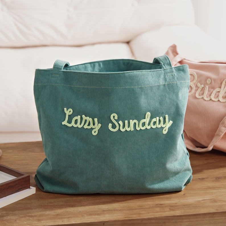 May include: A teal corduroy tote bag with the embroidered text "Lazy Sunday" in a light green script. The bag has two handles and is sitting on a wooden surface. A pink tote bag with the word "Bride" is partially visible.