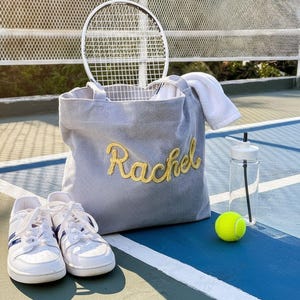 May include: A gray corduroy tote bag with the name "Rachel" embroidered in gold, a tennis racket, a white towel, a water bottle, a tennis ball, and white sneakers with blue stripes, all on a tennis court.
