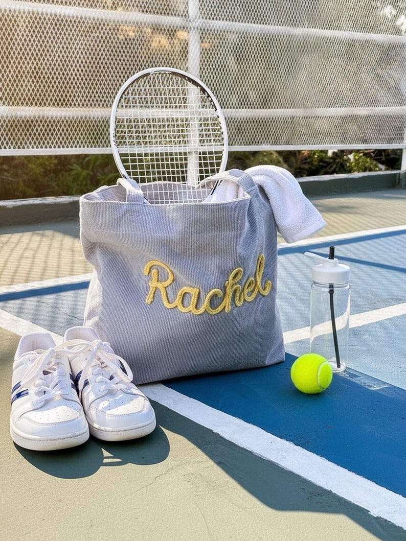May include: A gray corduroy tote bag with the name "Rachel" embroidered in gold, a tennis racket, a white towel, a water bottle, a tennis ball, and white sneakers with blue stripes, all on a tennis court.