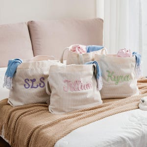 May include: Four beige canvas tote bags with embroidered names in purple, pink, and green. Each bag has a handle and is filled with items. Blue and white fringed scarves are draped over some of the bags.