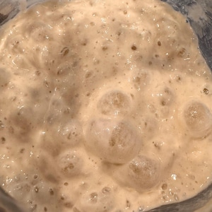 May include: Close-up of a light brown sourdough starter in a glass jar. The starter is bubbly and has a thick consistency.