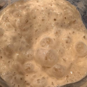 May include: Close-up of a light brown sourdough starter in a glass jar. The starter is bubbly and has a thick consistency.