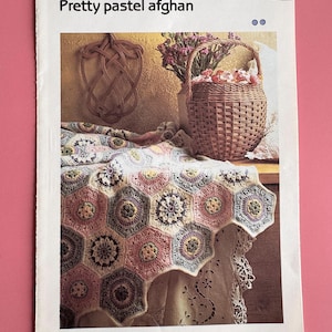 Puede incluir: Un patrón de crochet para una manta afgana de color pastel, con un diseño hexagonal. La manta está drapeada sobre una mesa con un mantel de encaje y una cesta de mimbre llena de flores.