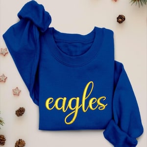 Puede incluir: Sudadera azul real con la palabra "eagles" bordada en dorado.