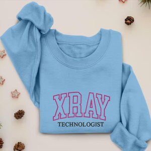 K&ouml;nnte beinhalten: Hellblauer Pullover mit dem Text "XRAY TECHNOLOGIST" in schwarzer und pinkfarbener Schrift.