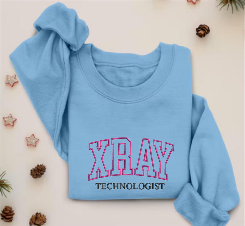 K&ouml;nnte beinhalten: Hellblauer Pullover mit dem Text "XRAY TECHNOLOGIST" in schwarzer und pinkfarbener Schrift.