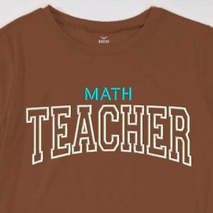 Op de afbeelding: Een bruin t-shirt met de geborduurde tekst "MATH TEACHER" in wit met turquoise accenten. Het shirt heeft een ronde hals en korte mouwen. Het ontwerp is gecentreerd op de borst.