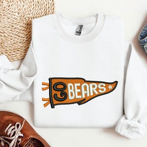 May include: White sweatshirt with an embroidered orange pennant design. The pennant reads "GO BEARS" in white lettering with a black outline. The sweatshirt is paired with blue jeans and brown shoes.