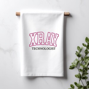 K&ouml;nnte beinhalten: Wei&szlig;es K&uuml;chentuch mit dem Text "XRAY TECHNOLOGIST" in einer pinkfarbenen und wei&szlig;en College-Schrift.
