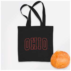 May include: Black canvas tote bag with red outline of the word "OHIO" in a collegiate font. A small orange pumpkin is in the lower right corner of the image.